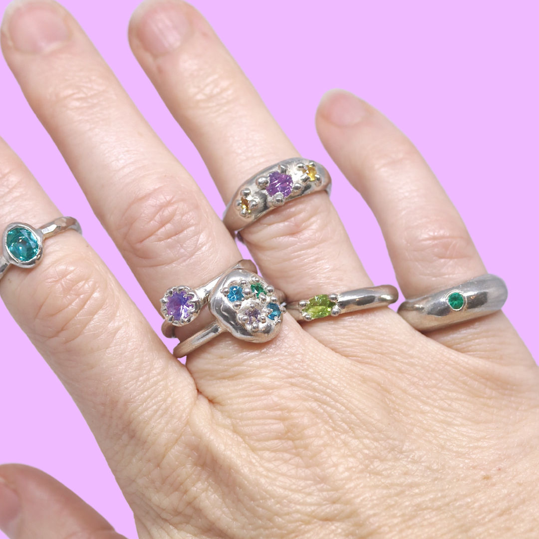 Hand wearing multiple silver rings with colorful gemstones on a pink background.