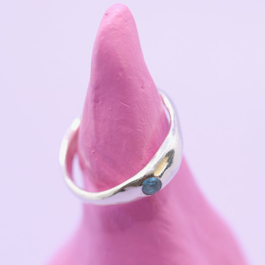 Silver ring with a blue gemstone on a pink mannequin finger against a light purple background