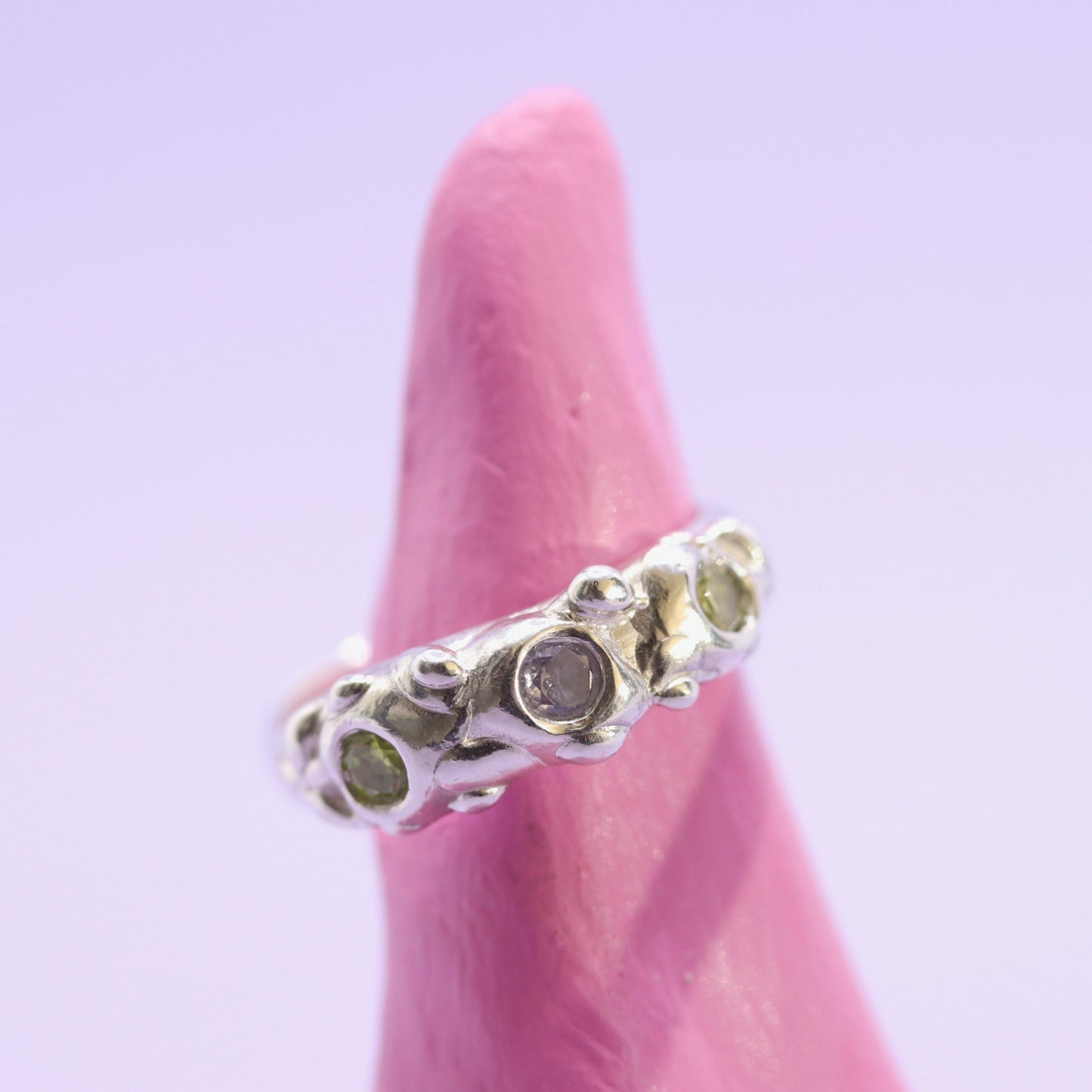 Silver ring with green gemstones on a pink finger against a light purple background