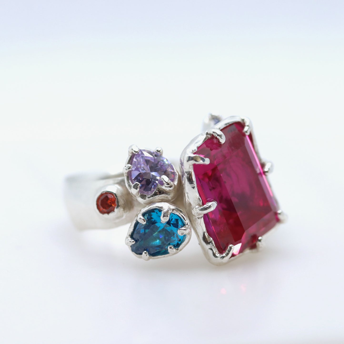 A silver ring featuring a large rectangular pink gemstone, a heart-shaped blue gemstone, a heart-shaped purple gemstone, and a small round red gemstone, set against a plain white background.