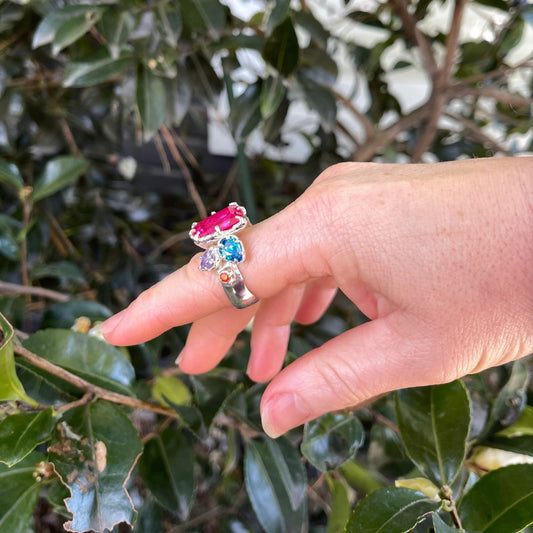 A hand wearing a silver ring with a large, colorful floral cluster featuring a prominent pink gemstone and a blue gemstone, set against a background of green leaves.