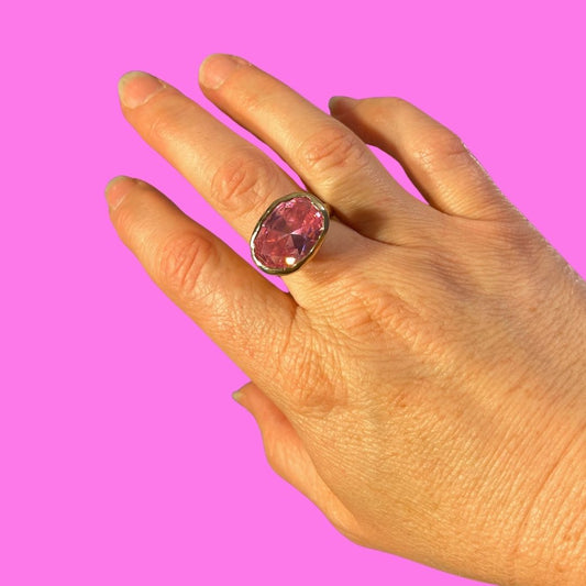 A hand wearing a vintage-style gold ring with a large oval pink gemstone, set against a solid bright pink background.