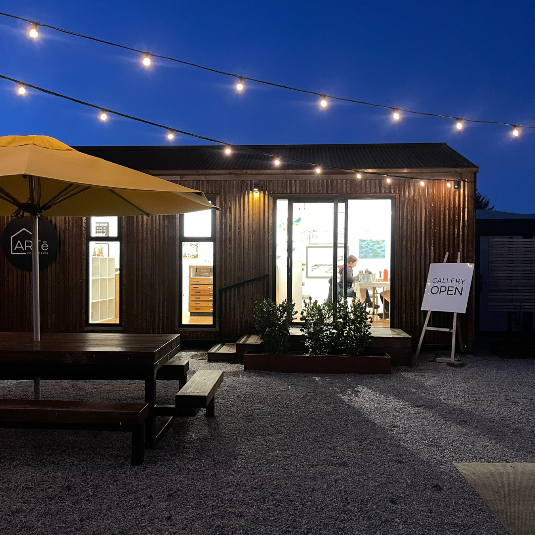 A modern wood-clad gallery space at dusk with string lights overhead, an outdoor patio umbrella, picnic table, and a sign reading 'GALLERY OPEN' on a gravel courtyard.