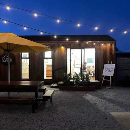 A modern wood-clad gallery space at dusk with string lights overhead, an outdoor patio umbrella, picnic table, and a sign reading 'GALLERY OPEN' on a gravel courtyard.