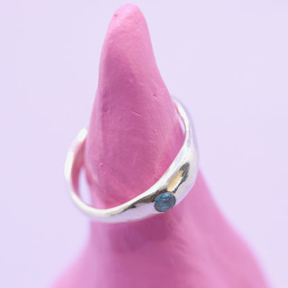 Silver ring with a blue gemstone on a pink mannequin finger against a light purple background