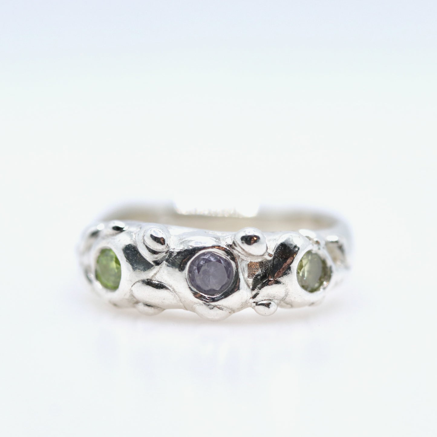 Silver ring with a central purple stone and two green stones on a white background