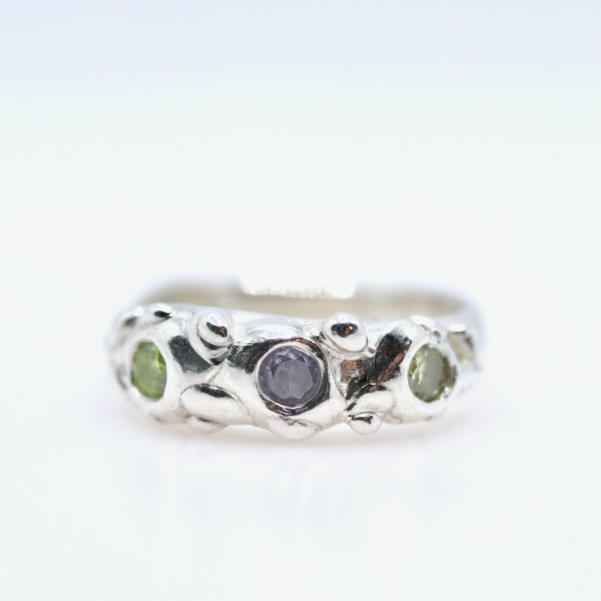 Silver ring with a central purple stone and two green stones on a white background