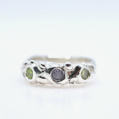 Silver ring with a central purple stone and two green stones on a white background