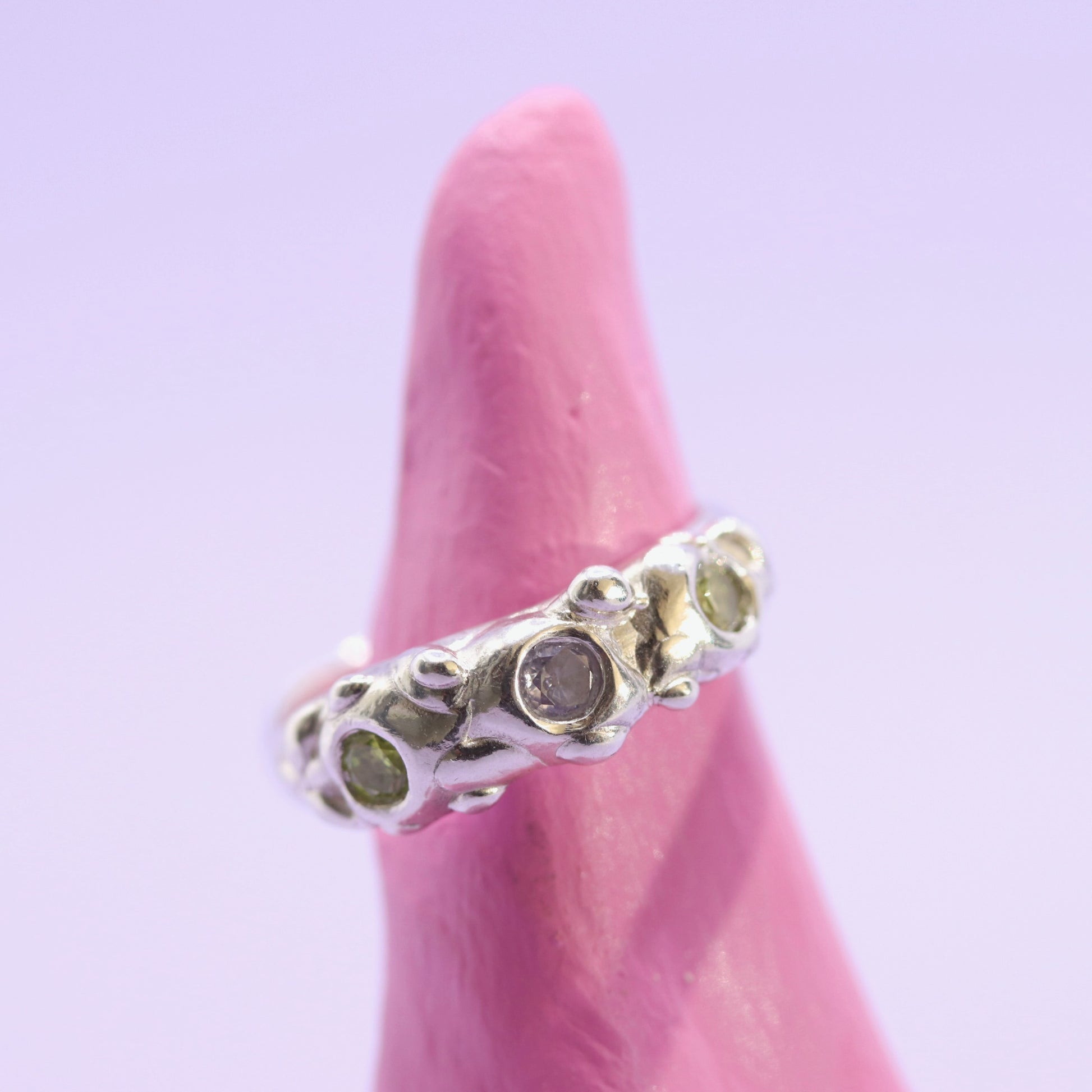 Silver ring with green gemstones on a pink finger against a light purple background