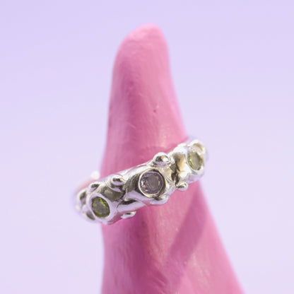 Silver ring with green gemstones on a pink finger against a light purple background