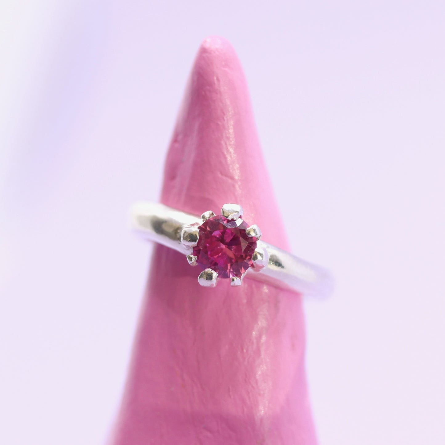 Silver ring with a pink gemstone on a pink stand against a light purple background