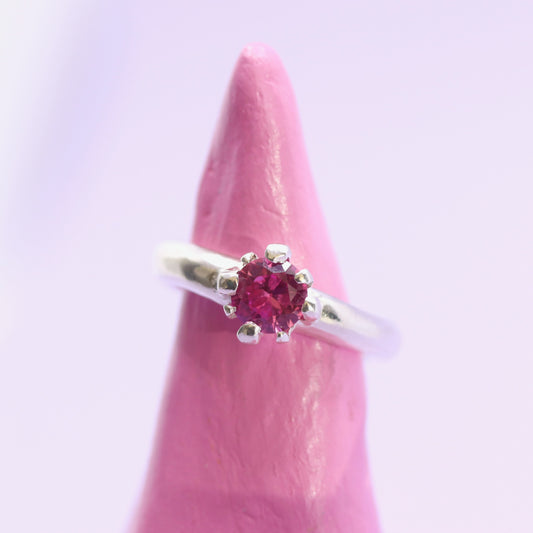 Silver ring with a pink gemstone on a pink stand against a light purple background