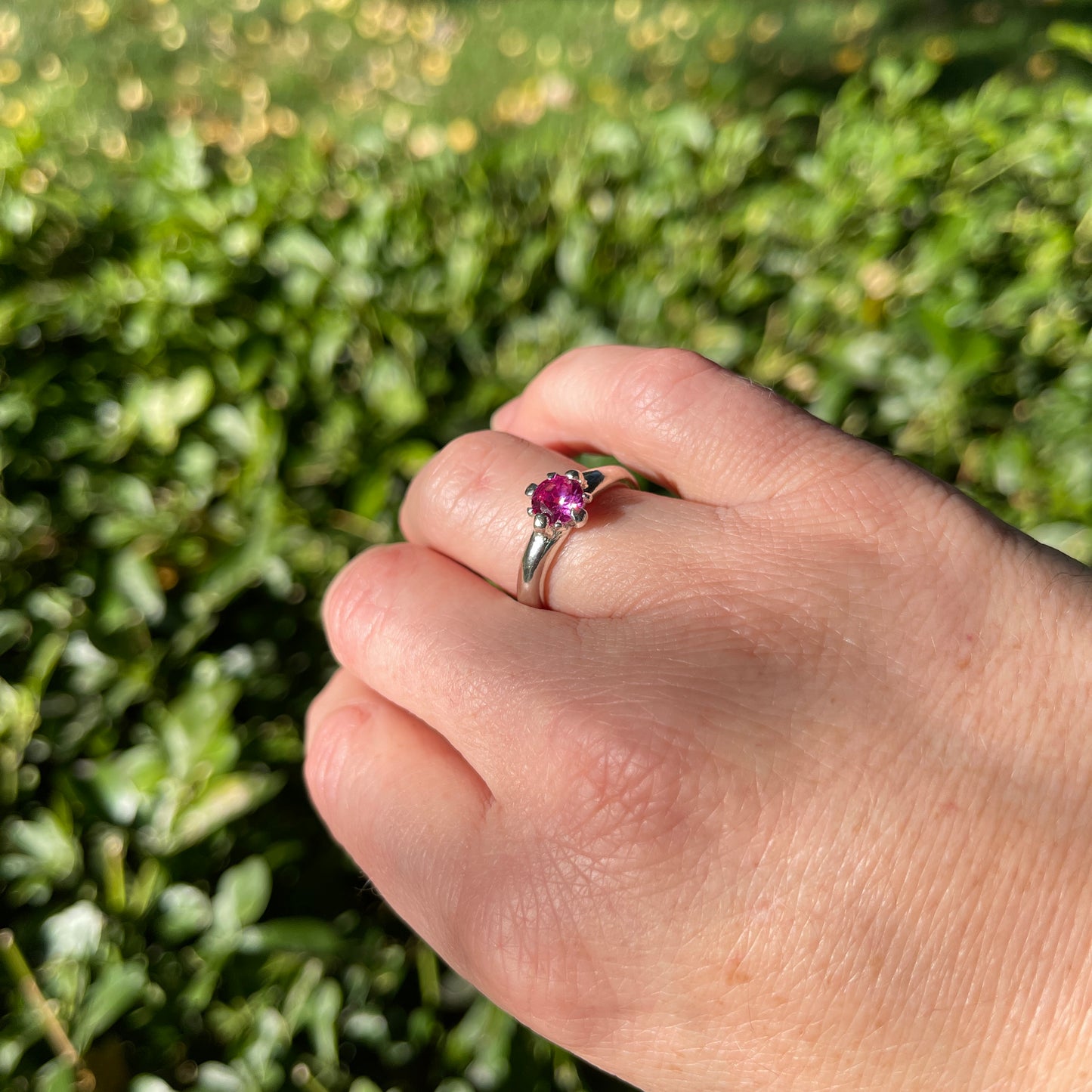 Hand wearing a ring with a pink gemstone against a green leafy background