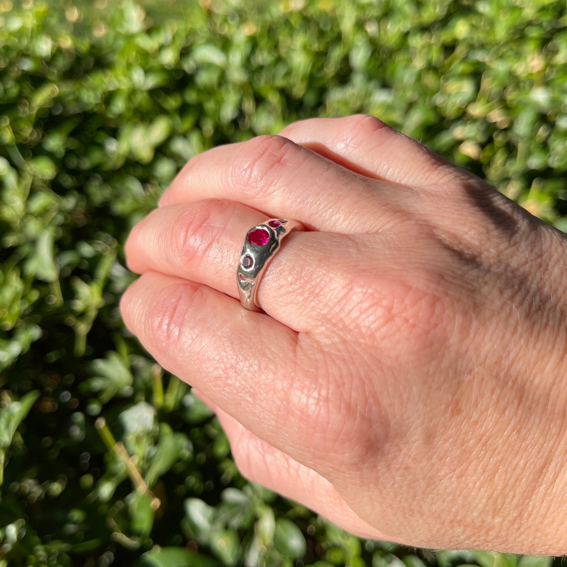 Hand wearing a silver ring with a ruby pink gemstone against a green leafy background