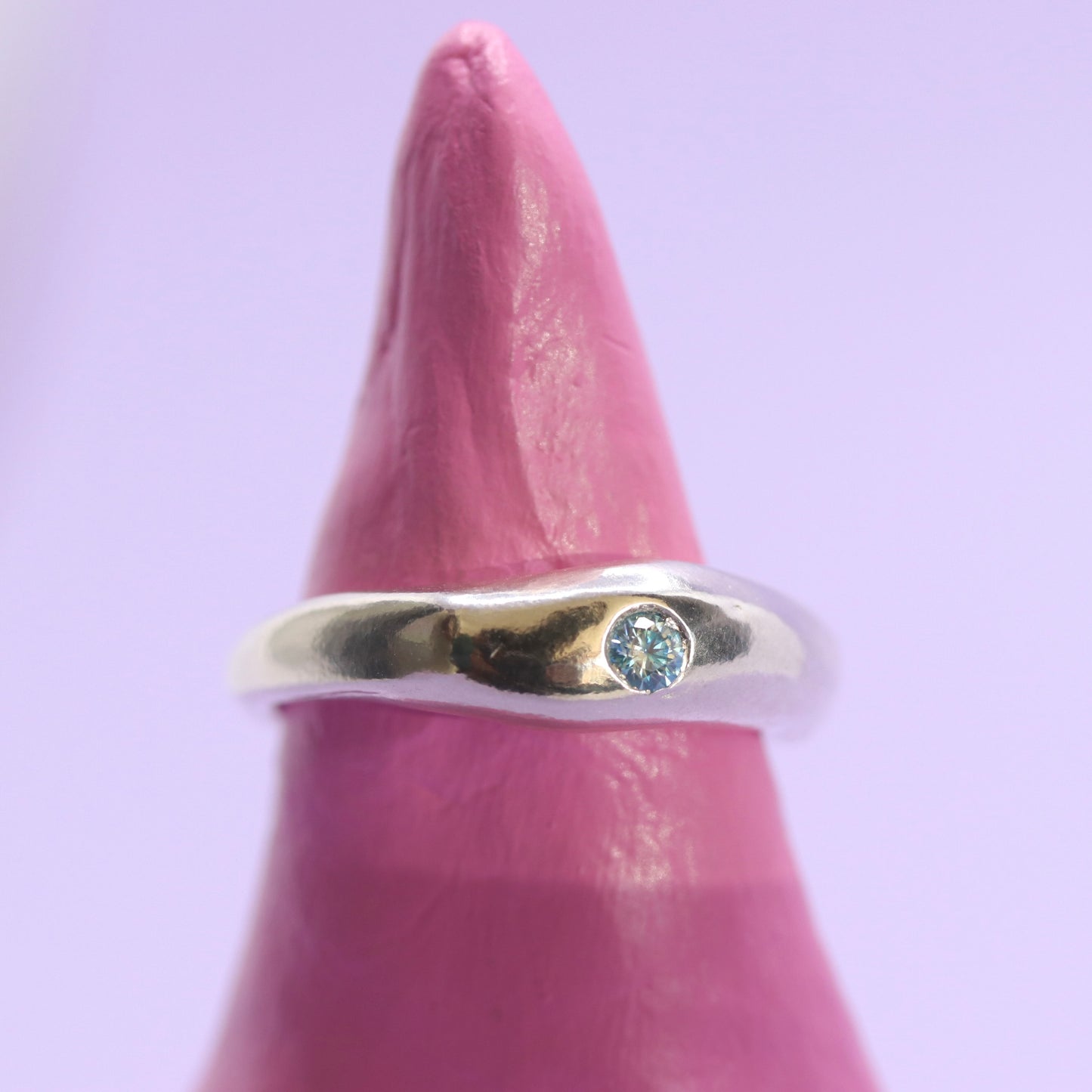Silver ring with a blue gemstone on a pink finger against a light purple background
