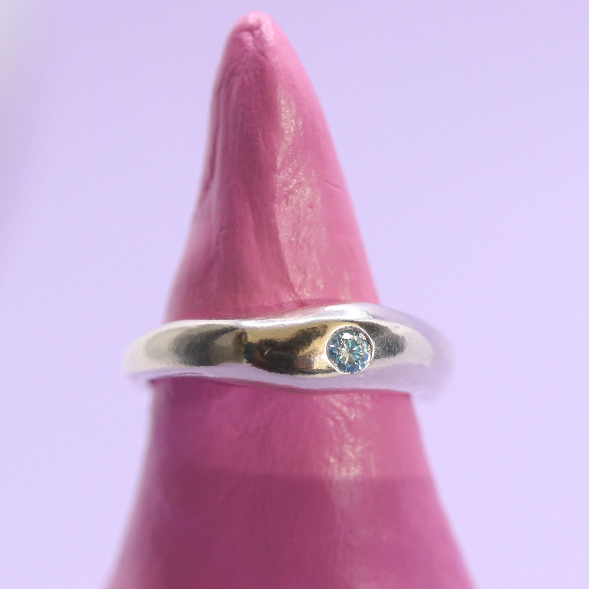 Silver ring with a blue gemstone on a pink finger against a light purple background
