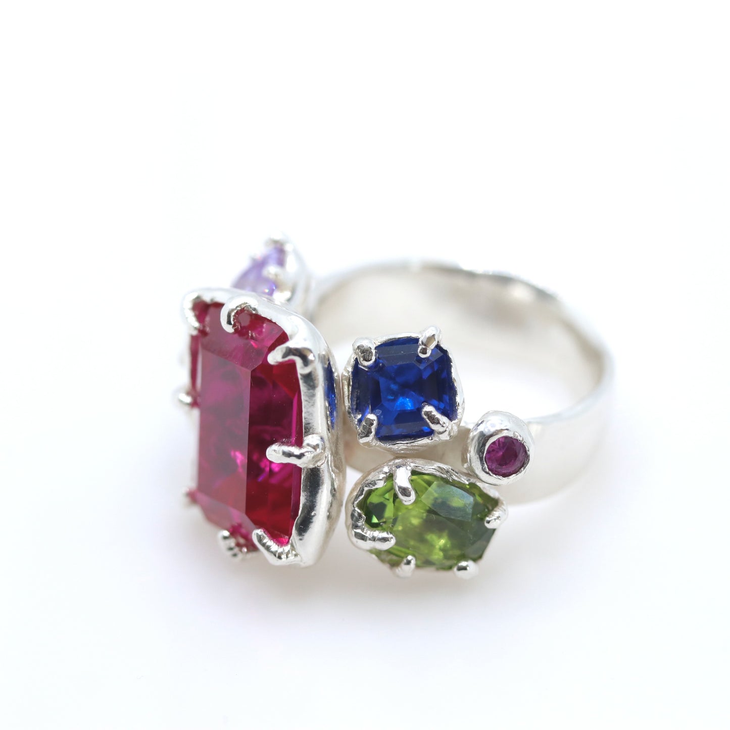 A silver ring featuring multiple colorful gemstones, including a large rectangular pinkish-red stone, a square blue stone, a green stone, and smaller purple and magenta stones, set against a plain white background.
