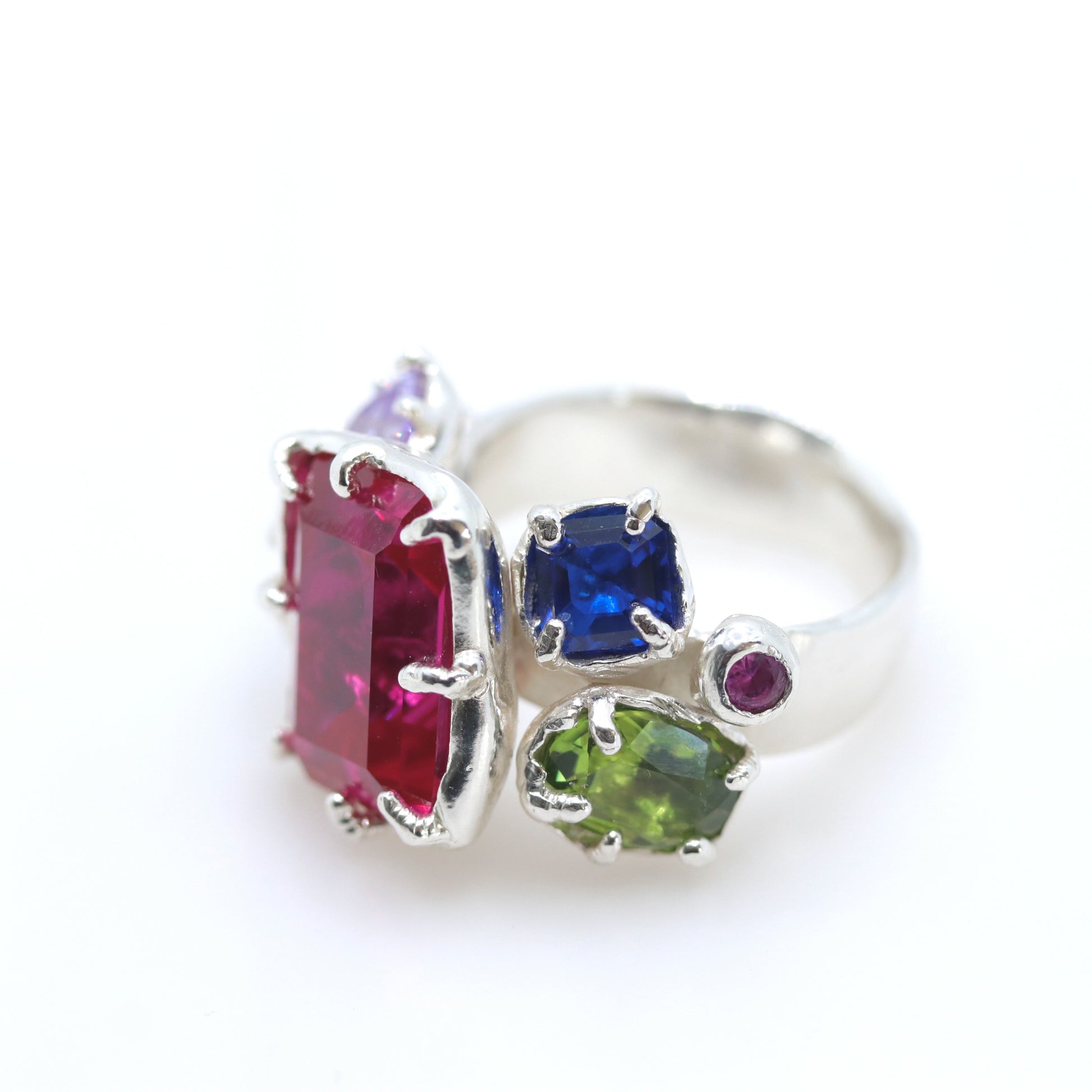 A silver ring featuring multiple colorful gemstones, including a large rectangular pinkish-red stone, a square blue stone, a green stone, and smaller purple and magenta stones, set against a plain white background.