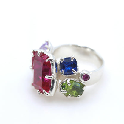 A silver ring featuring multiple colorful gemstones, including a large rectangular pinkish-red stone, a square blue stone, a green stone, and smaller purple and magenta stones, set against a plain white background.