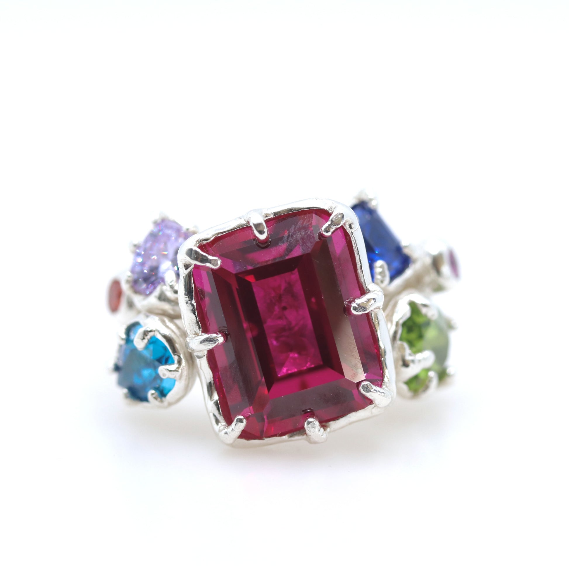 A silver ring featuring a large rectangular emerald-cut pinkish-red center stone, accented by smaller round-cut purple, blue, green, and dark blue stones on a white background.