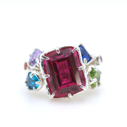 A silver ring featuring a large rectangular emerald-cut pinkish-red center stone, accented by smaller round-cut purple, blue, green, and dark blue stones on a white background.
