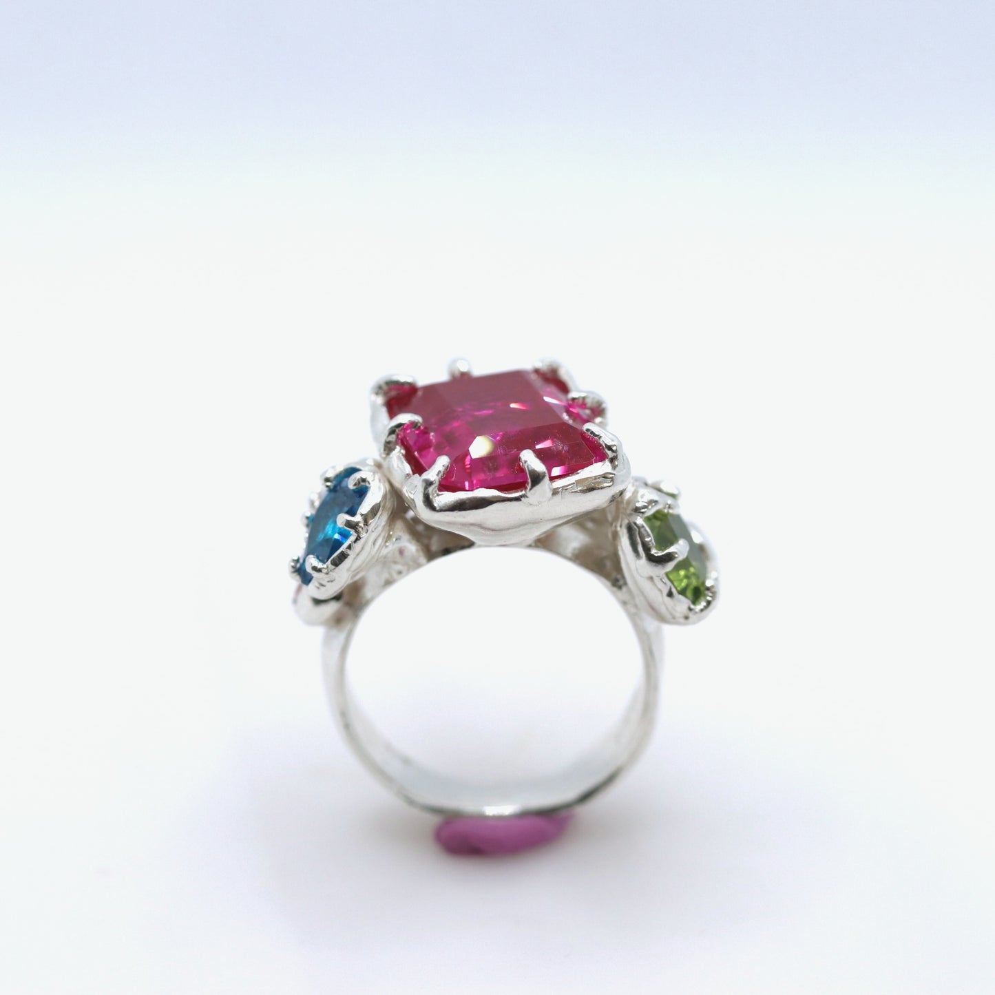 A silver ring featuring a large rectangular pink gemstone in the center, with smaller blue and green gemstones on either side, set against a plain white background.