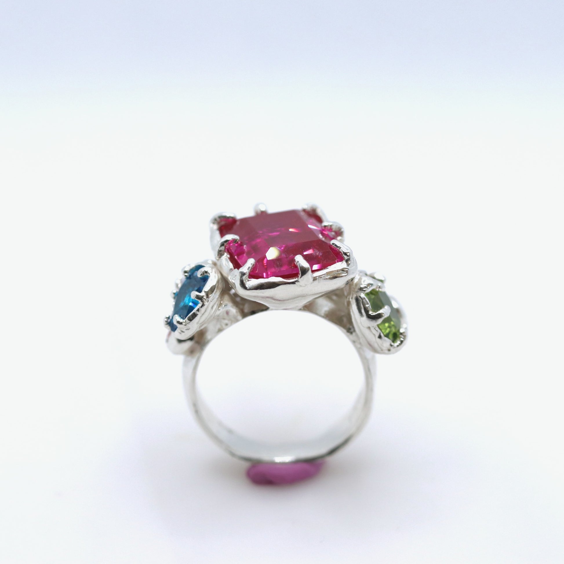 A silver ring featuring a large rectangular pink gemstone in the center, with smaller blue and green gemstones on either side, set against a plain white background.