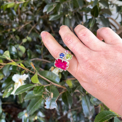 A hand wearing a silver ring with a large rectangular pink gemstone, a smaller square blue gemstone, and a round light green gemstone, set against a background of green leaves and white flowers.