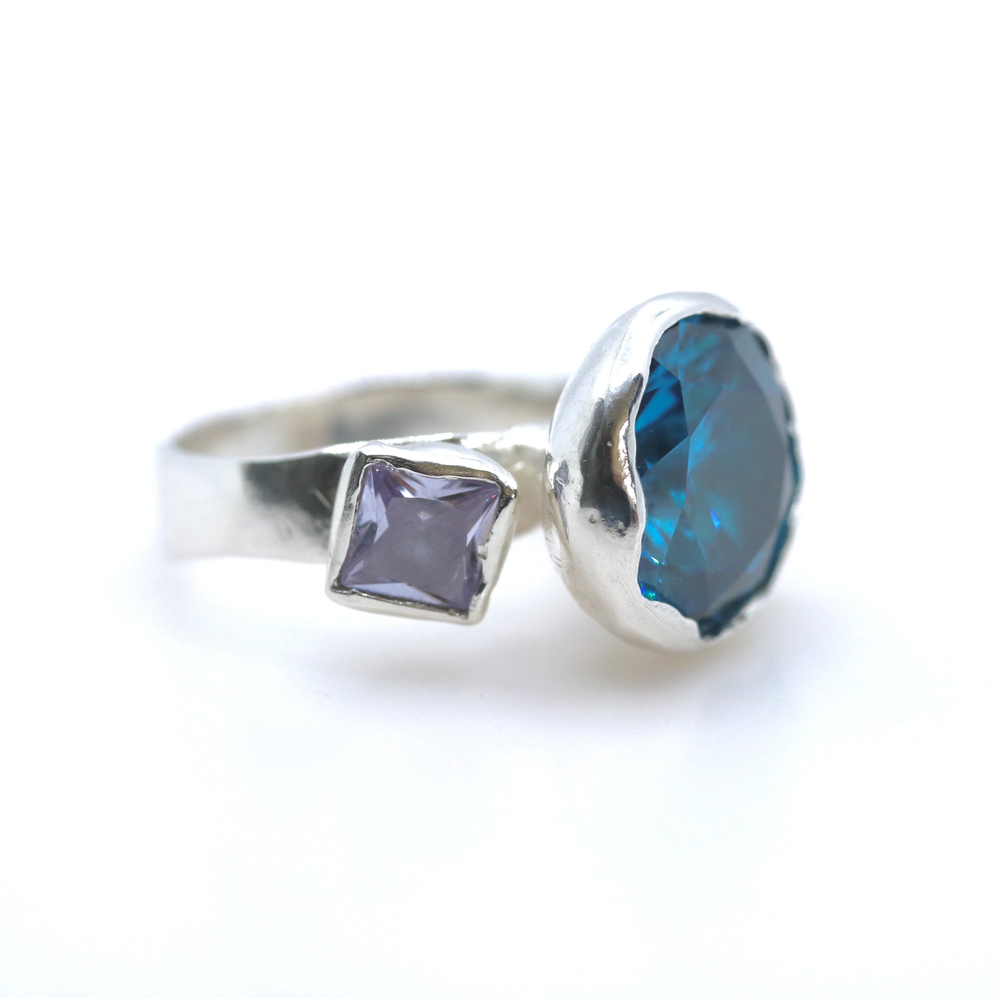 A silver ring featuring a large, irregular-cut deep blue faceted gemstone and a smaller square-shaped lavender-purple gemstone, set against a plain white background.