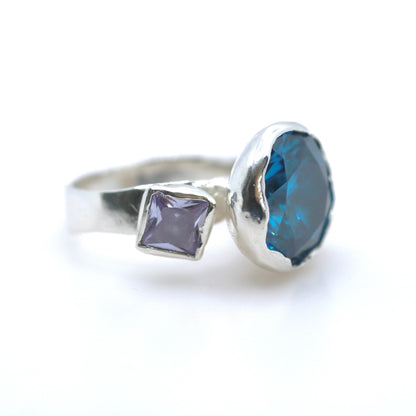 A silver ring featuring a large, irregular-cut deep blue faceted gemstone and a smaller square-shaped lavender-purple gemstone, set against a plain white background.