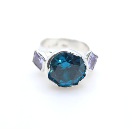 A silver ring featuring a large, faceted dark blue heart-shaped gemstone in the center, flanked by two small, rectangular light purple gemstones on a white background.