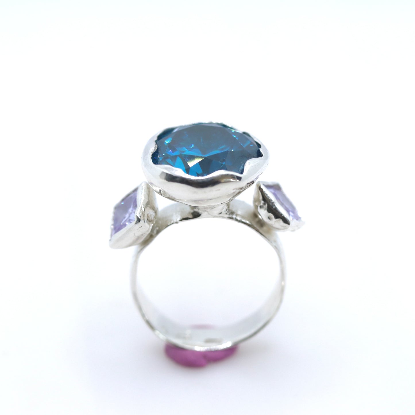 A silver ring featuring a large, scalloped-edge blue gemstone in the center, flanked by two smaller light purple rectangular gemstones on the shoulders, set against a plain white background.