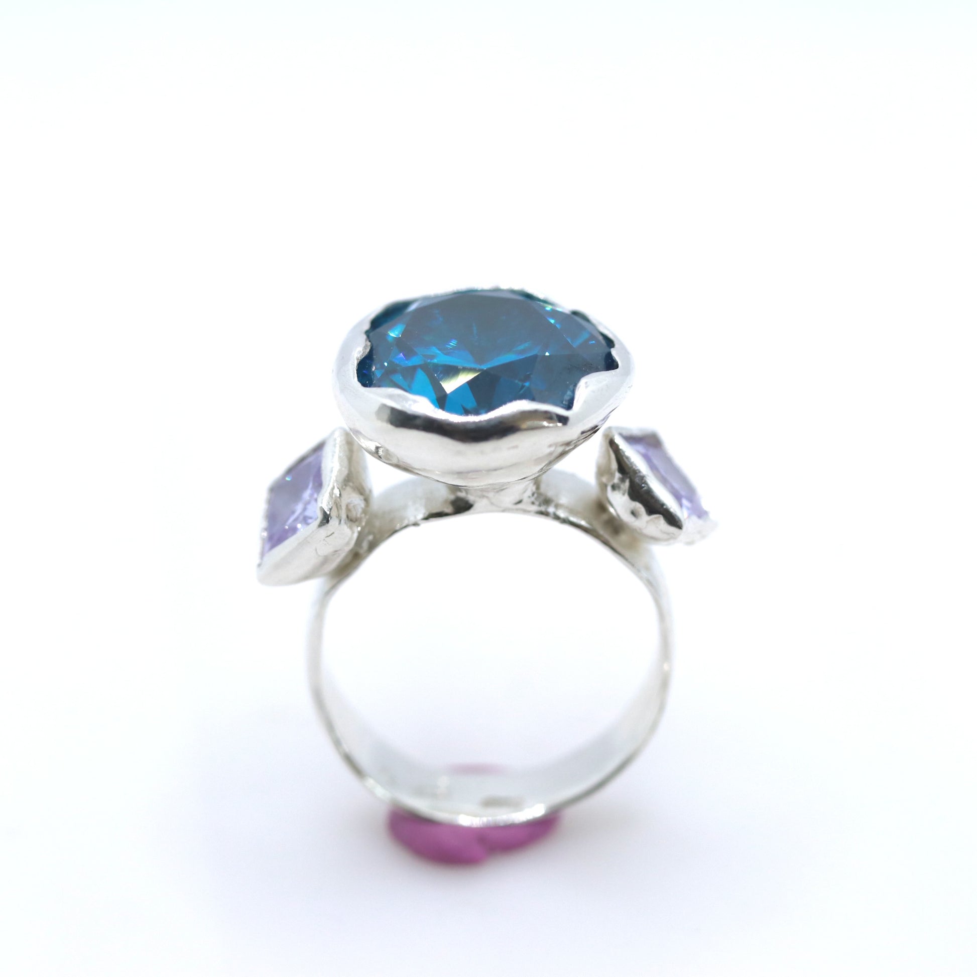 A silver ring featuring a large, scalloped-edge blue gemstone in the center, flanked by two smaller light purple rectangular gemstones on the shoulders, set against a plain white background.