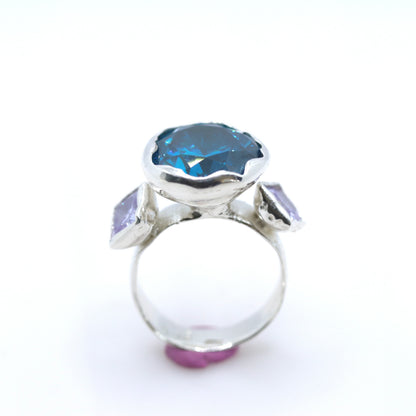 A silver ring featuring a large, scalloped-edge blue gemstone in the center, flanked by two smaller light purple rectangular gemstones on the shoulders, set against a plain white background.
