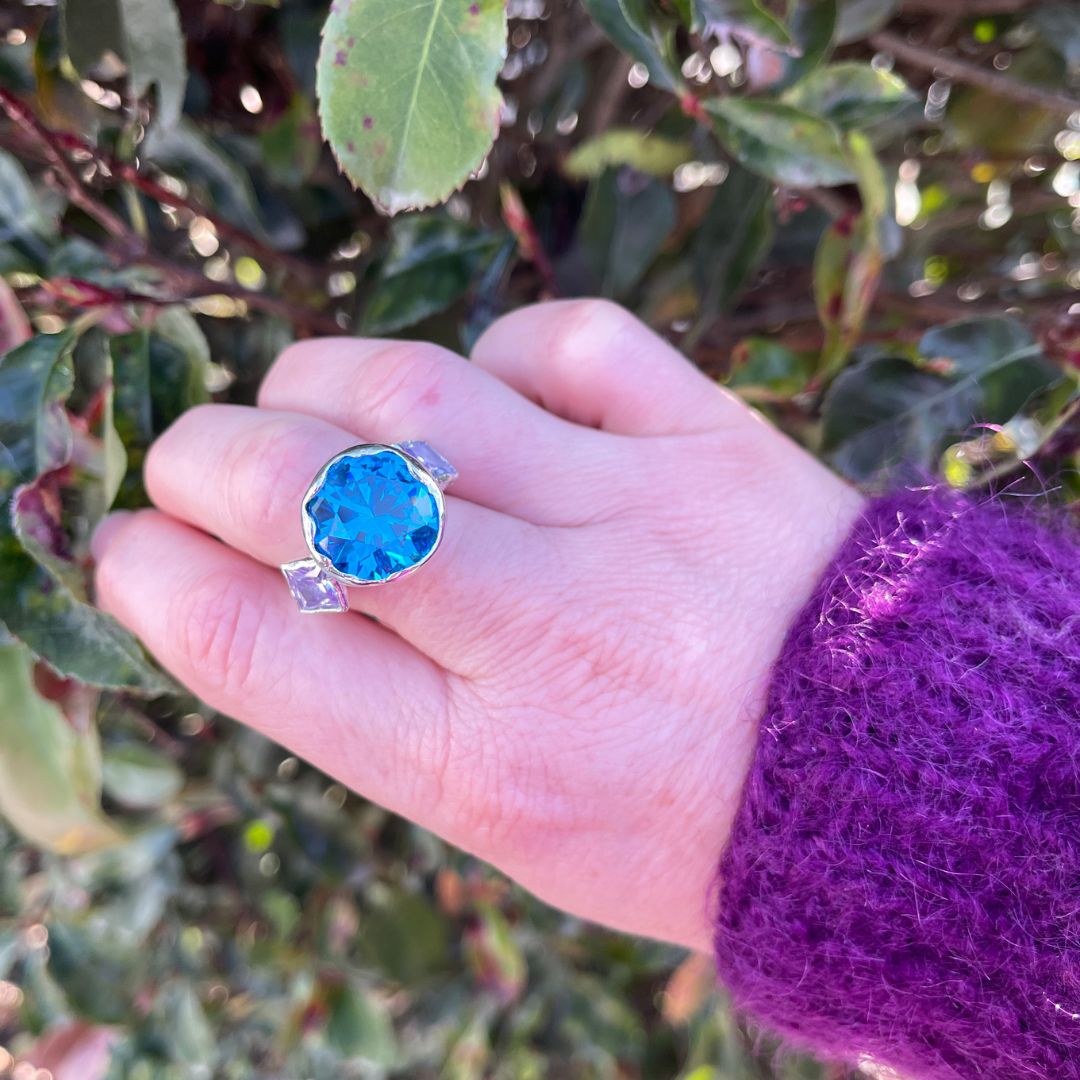 A hand wearing a silver ring with a large, scalloped oval-cut bright blue gemstone, set against a background of green leaves and a purple knit sleeve.