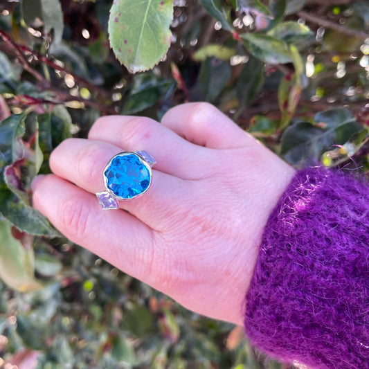 A hand wearing a silver ring with a large, scalloped oval-cut bright blue gemstone, set against a background of green leaves and a purple knit sleeve.
