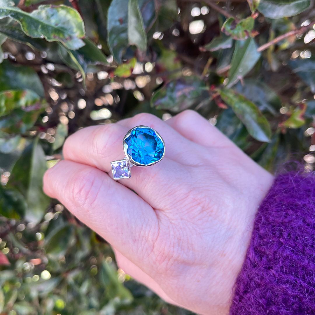 A close-up of a hand wearing a silver ring with a large, round, bright blue gemstone and a smaller square clear gemstone, set against a background of green foliage.
