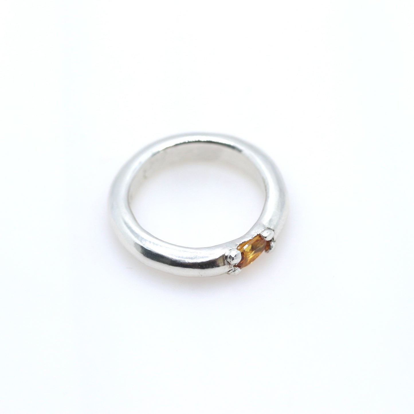 A polished silver ring with a small, round, amber-colored gemstone set on the front, photographed on a seamless white background.
