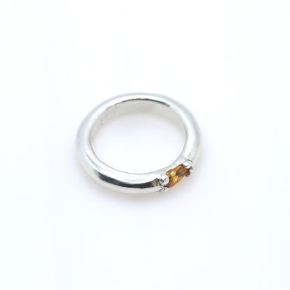 A polished silver ring with a small, round, amber-colored gemstone set on the front, photographed on a seamless white background.