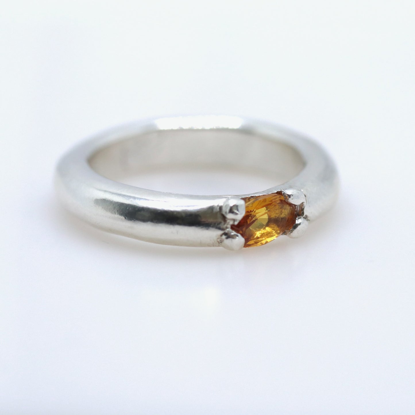 A silver band ring featuring a small, oval-cut amber gemstone in a prong setting, photographed on a clean white background.