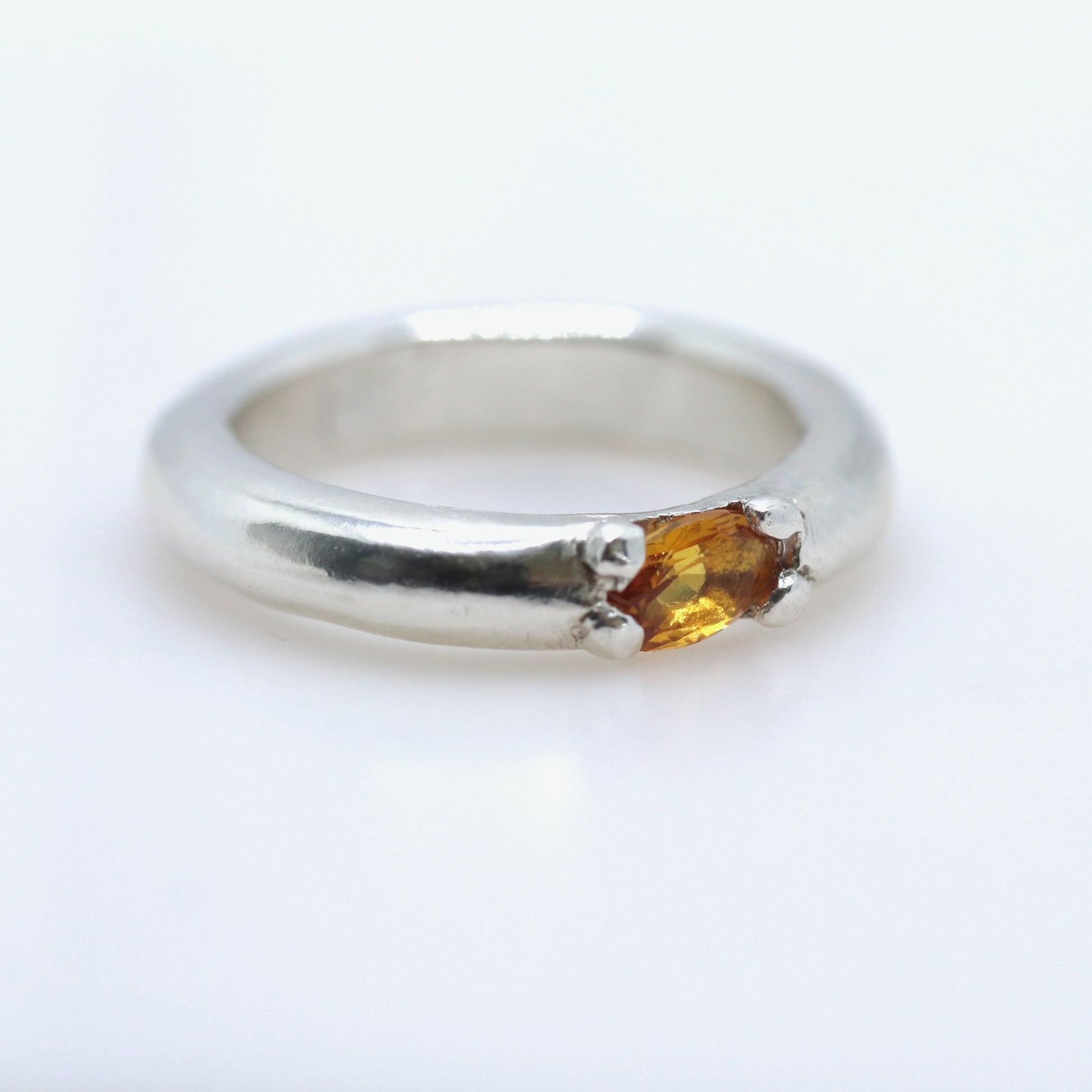 A silver band ring featuring a small, oval-cut amber gemstone in a prong setting, photographed on a clean white background.