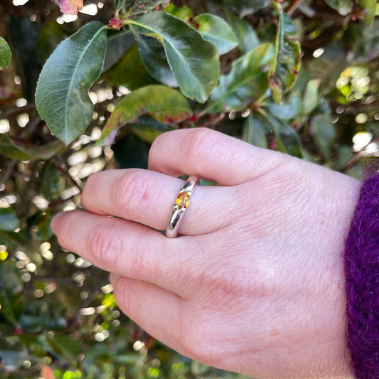 A close-up of a hand wearing a silver band ring with a small yellow gemstone, set against a background of green leaves and sunlight.