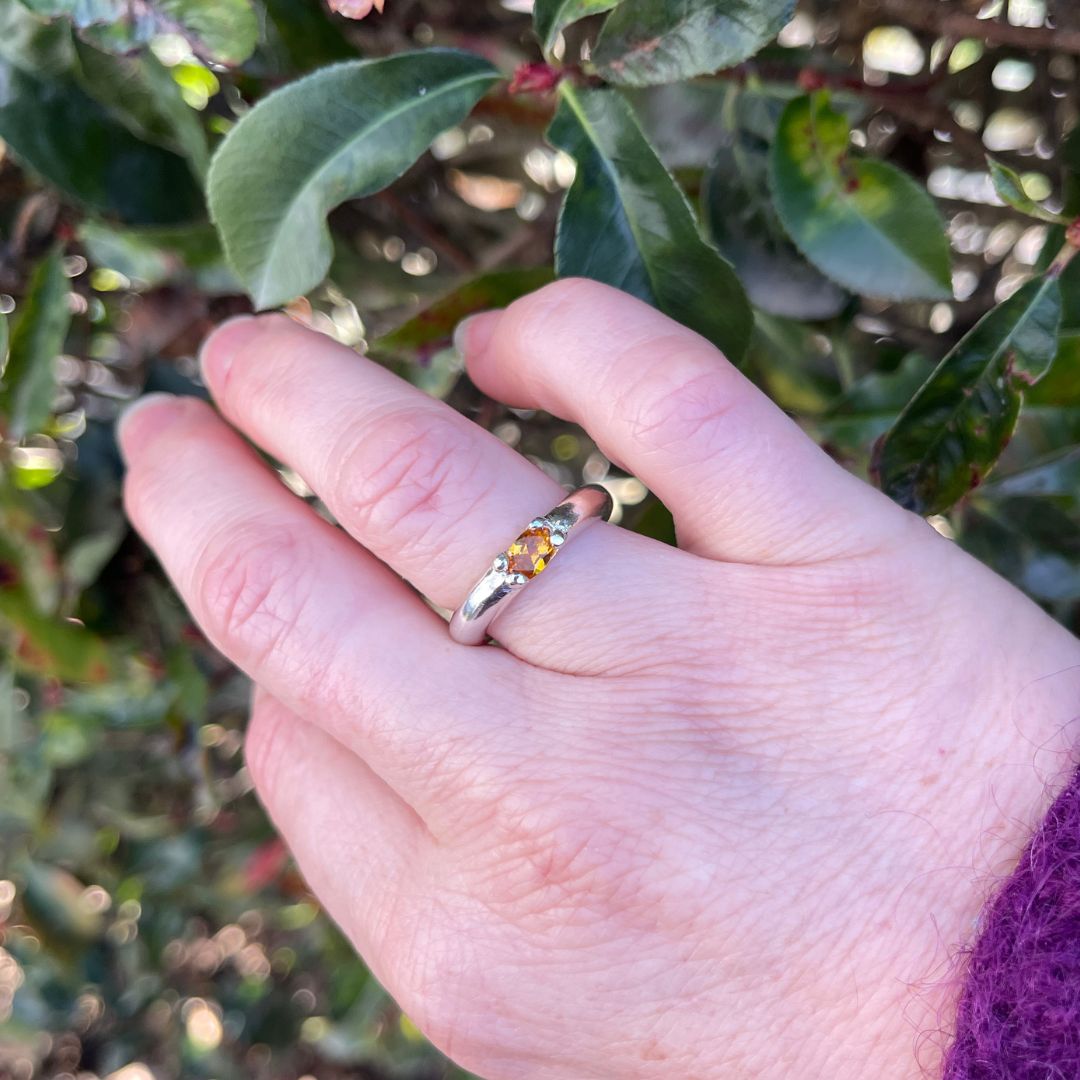A close-up of a hand wearing a silver ring with a small yellow gemstone, set against a blurred background of green leaves.