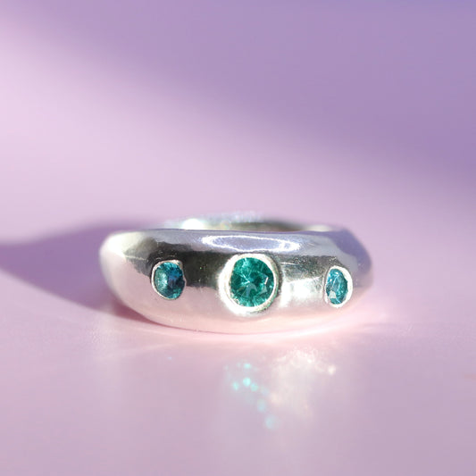 A silver ring with three round-cut emerald-cut green gemstones, displayed on a soft pink-lavender background.