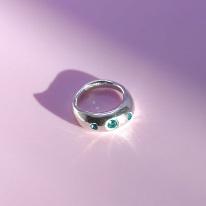 A polished silver ring featuring three small round green gemstones, photographed on a soft pink background with a bright light reflection and shadow.