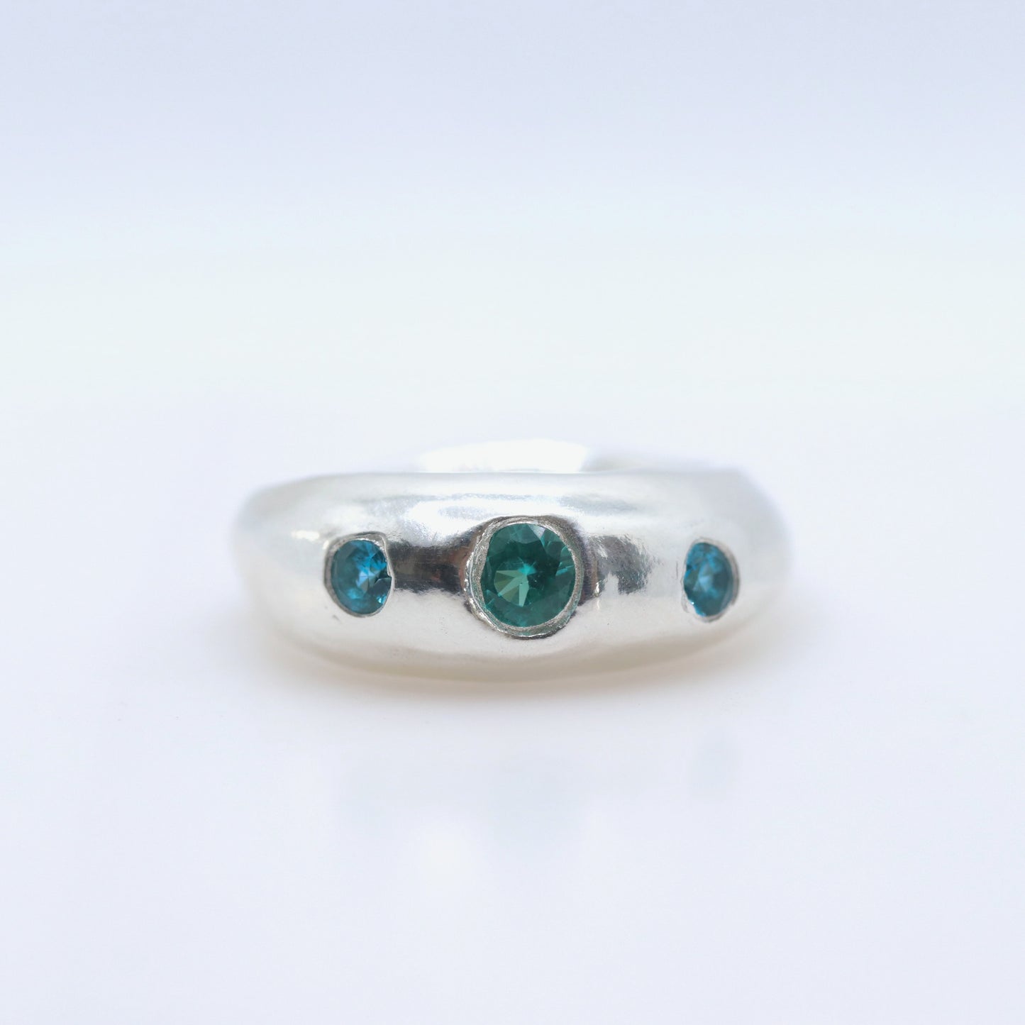 A silver band ring featuring three round-cut green gemstones, with one larger central stone and two smaller side stones, set against a plain white background.