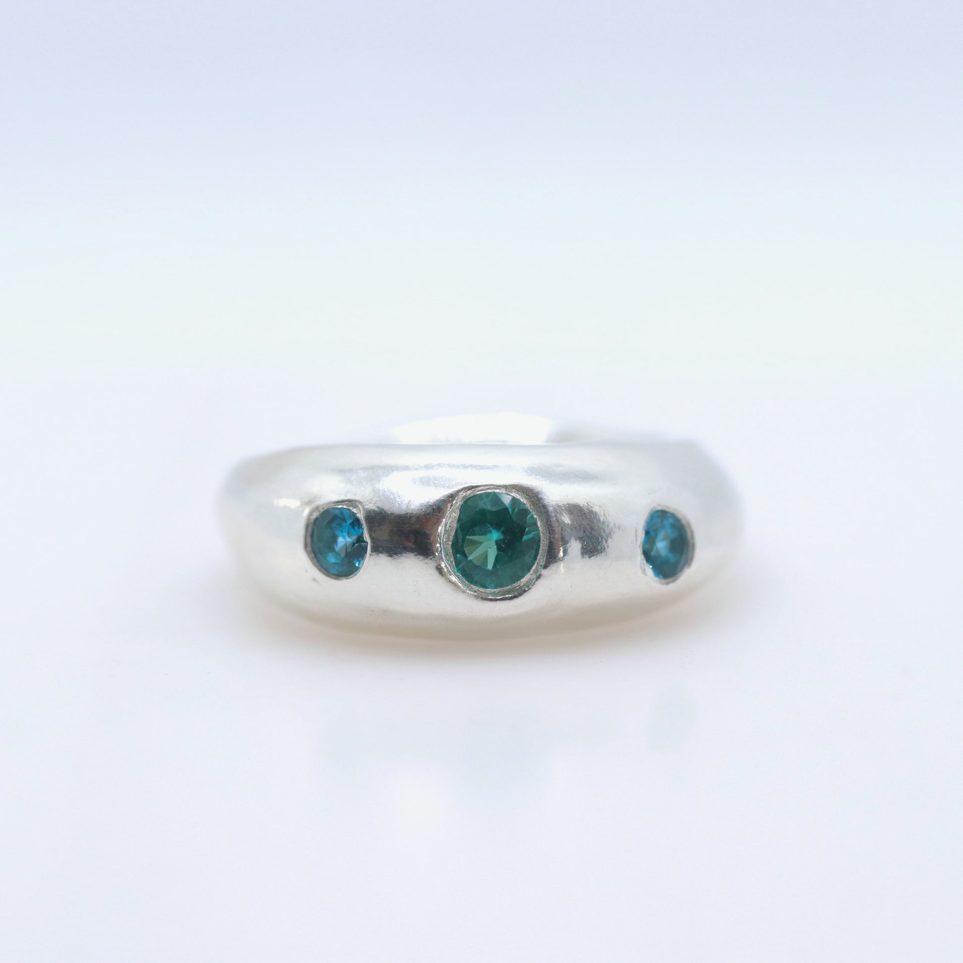 A silver band ring featuring three round-cut green gemstones, with one larger central stone and two smaller side stones, set against a plain white background.