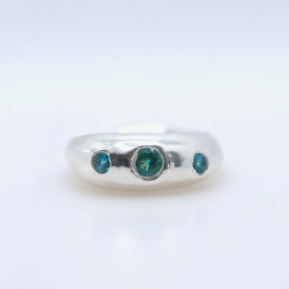 A silver band ring featuring three round-cut green gemstones, with one larger central stone and two smaller side stones, set against a plain white background.