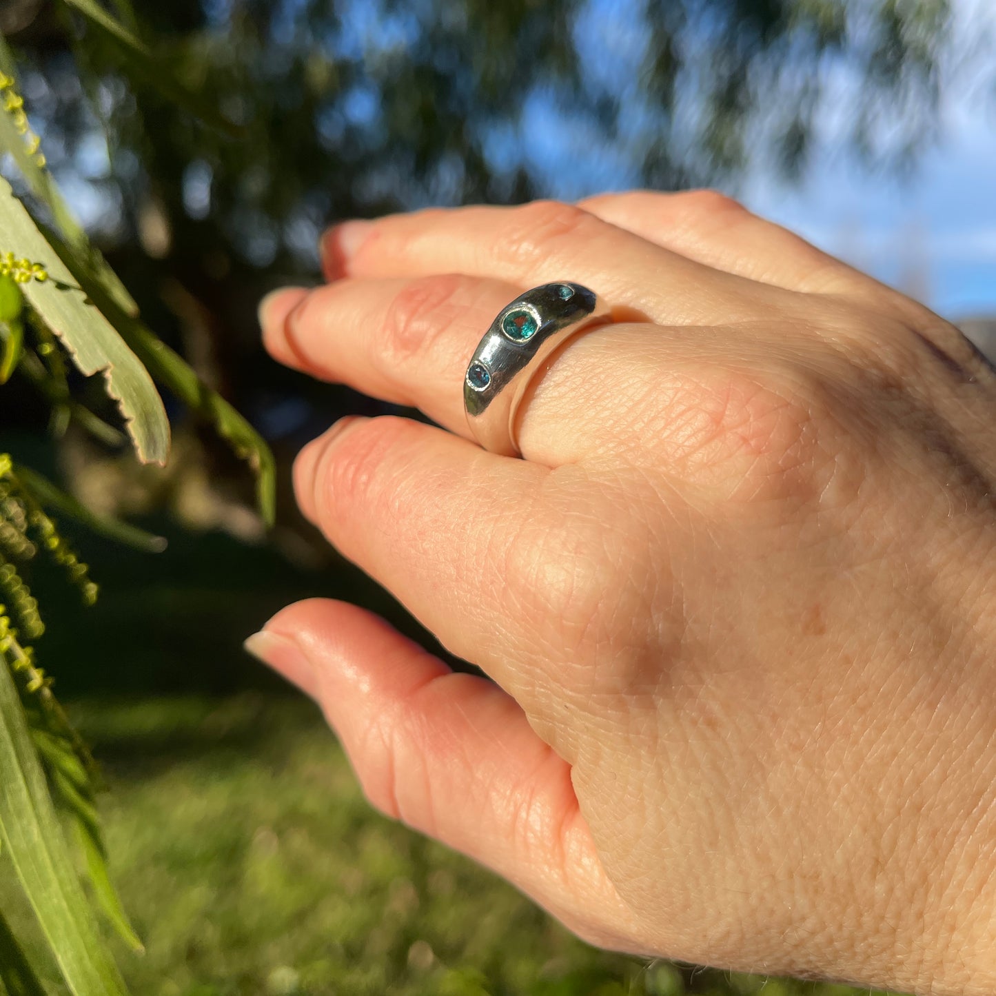 A close-up of a hand wearing a silver ring with two small round turquoise-blue stones, photographed outdoors with blurred green leaves and blue sky in the background.
