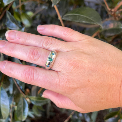 A gold band ring with three small green and blue gemstones, shown worn on a person's hand against a background of green leaves.